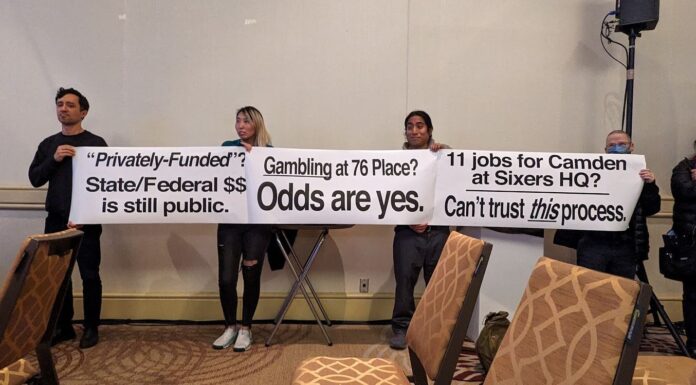 Protesters stand in the back of the room during a community meeting holding up signs asking pointed questions about gambling, public funding for a private arena and the promise of jobs