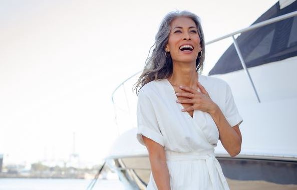 Nina Cash in a white dress stands in front of a yacht smiling broadly