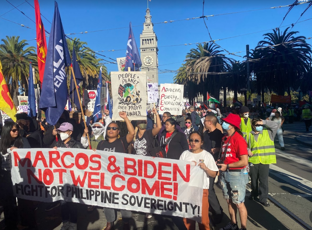 Marchers carry a sign that reads "Marcos and Biden not welcomed."