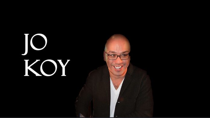 Jo Koy in a t-shirt and sports coat over a dark background and his name in white lettering