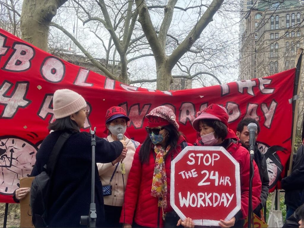 Hunger strikers introduce themselves onstage. One holds a sign reading "Stop the 24hr workday."