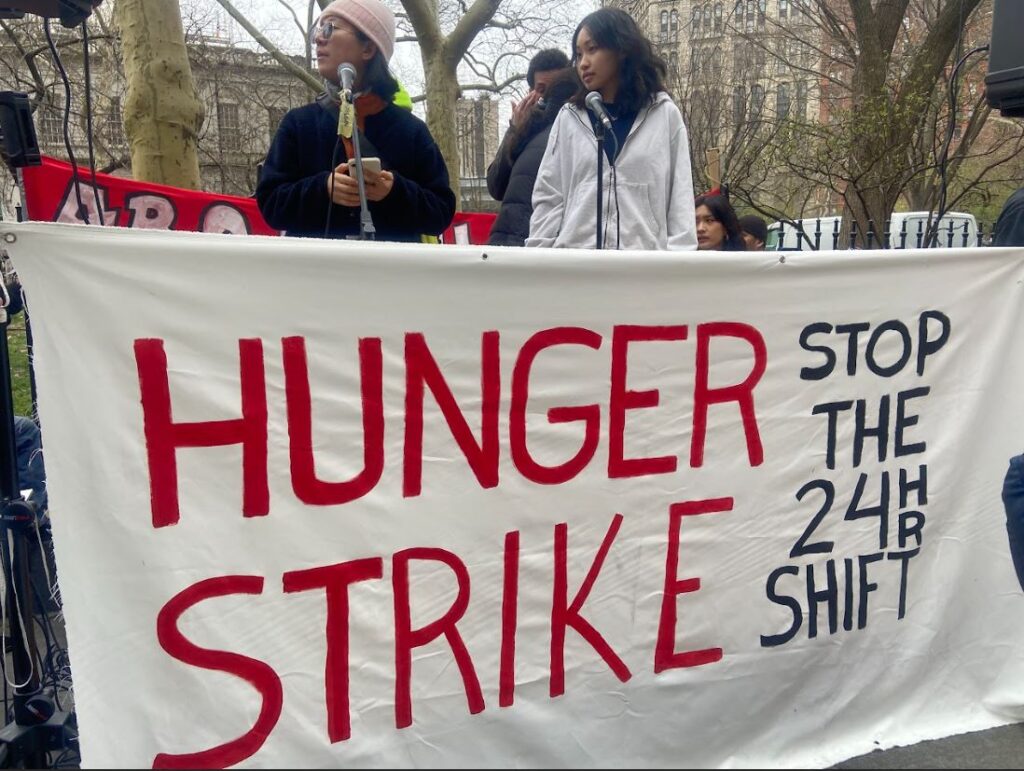 Two MCs for the rally stand on stage. A banner in front of them reads "Hunger Strike Stop the 24HR Shift"