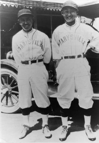 Two men in baseball uniforms posed before an old car.