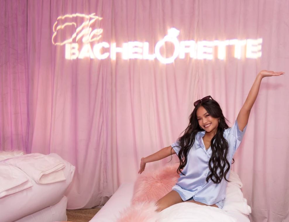 Jenn Tran poses on a bed in a nightgown in front of a Bachelorette neon sign