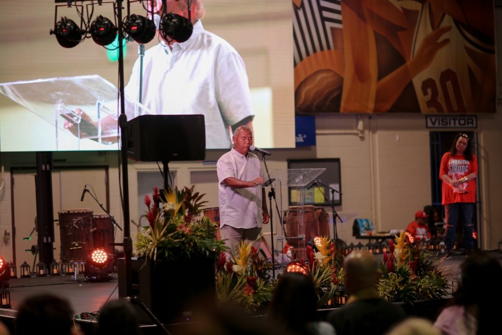 An elderly man in a white shirt speaks in front of the Lahaina Civic Center auditorium.