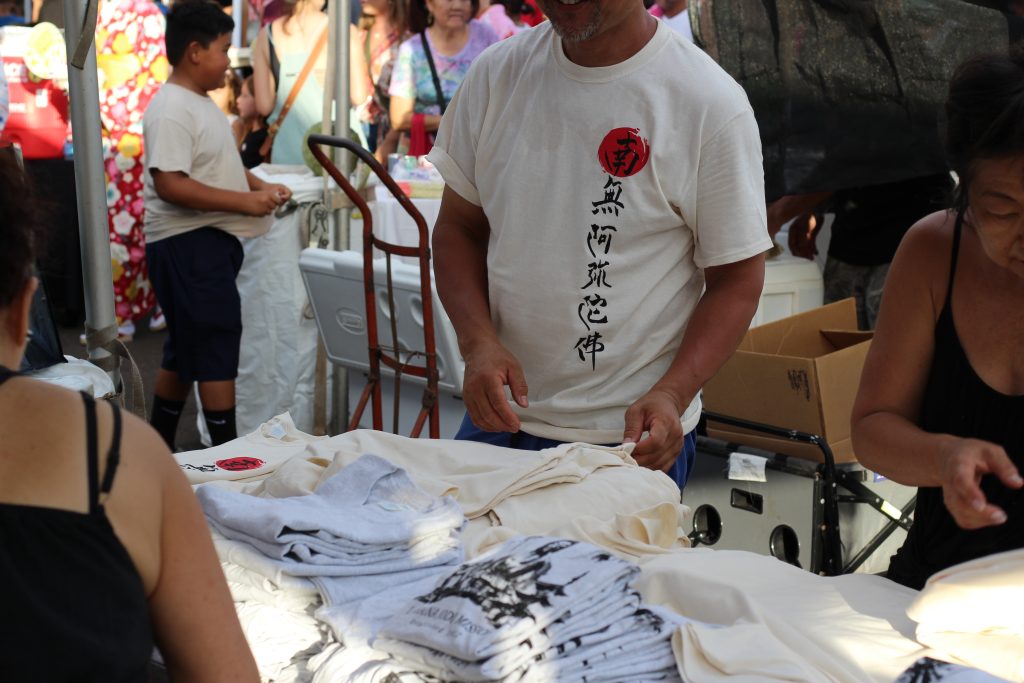 A man stands at a table selling t-shirts with Japanese characters.