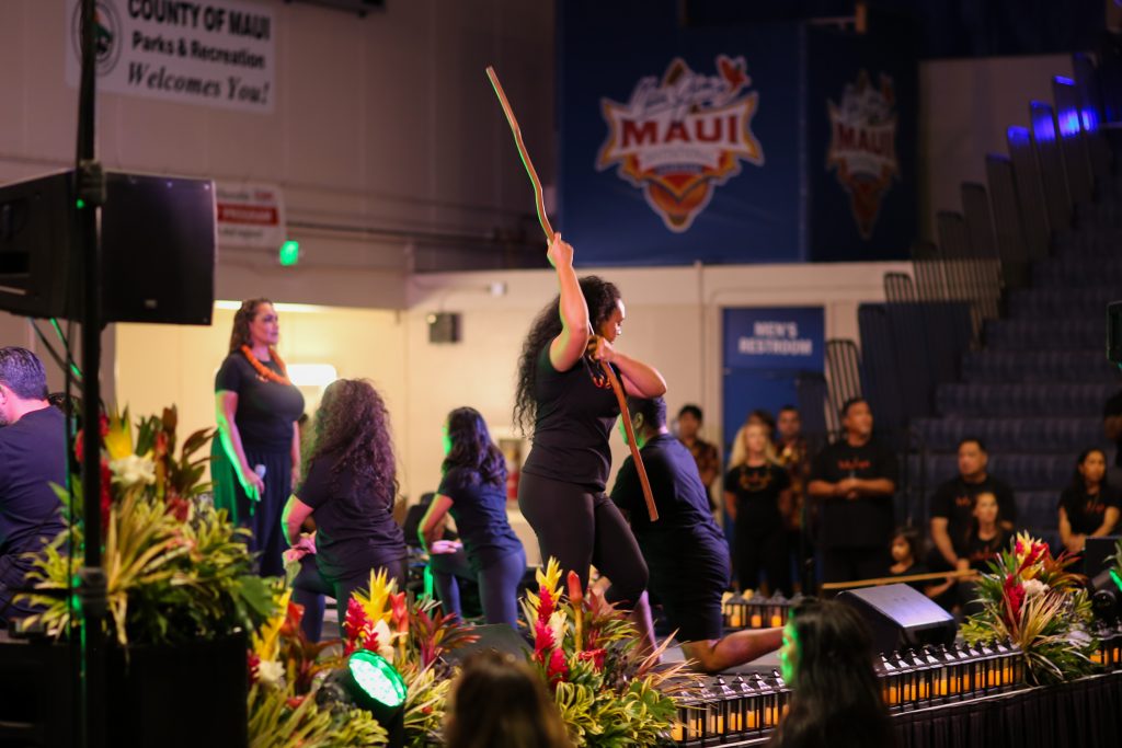 A dancer dances with a tall wooden stick at the Lahaina Civic Center gymnasium.