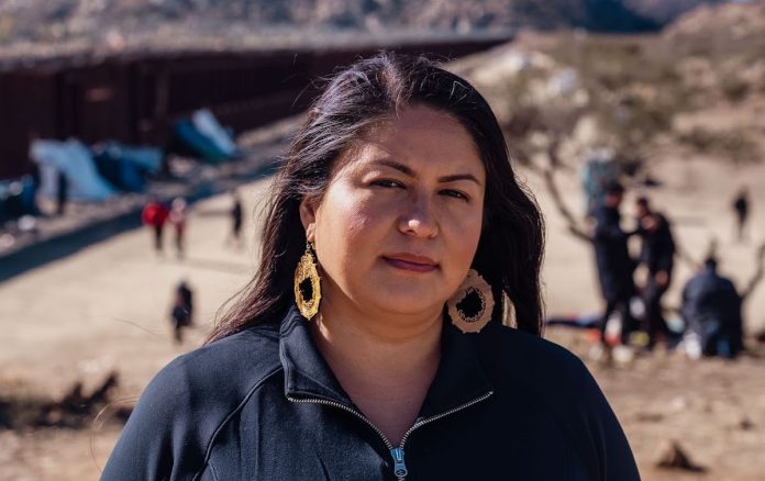 Lilian Serrano in front of the border wall at the U.S. Mexican border with asylum seekers, refugees and undocumented immigrants behind her