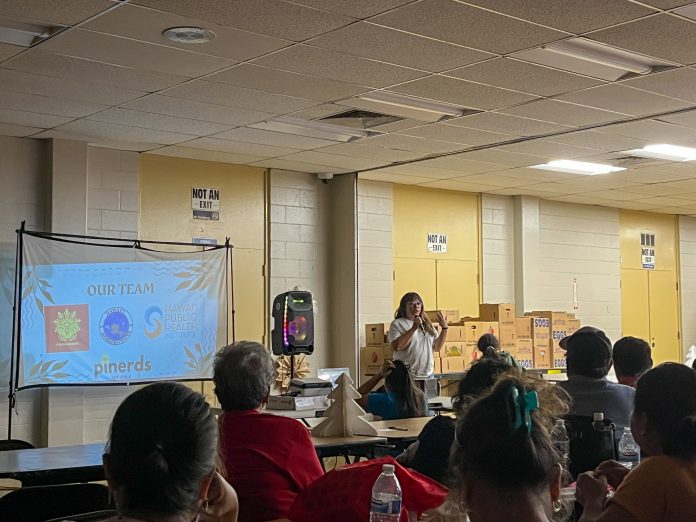 A woman stands in front of a screen with boxes of food from the food pantry behind her. Filipino elders sit in the audience and listen.