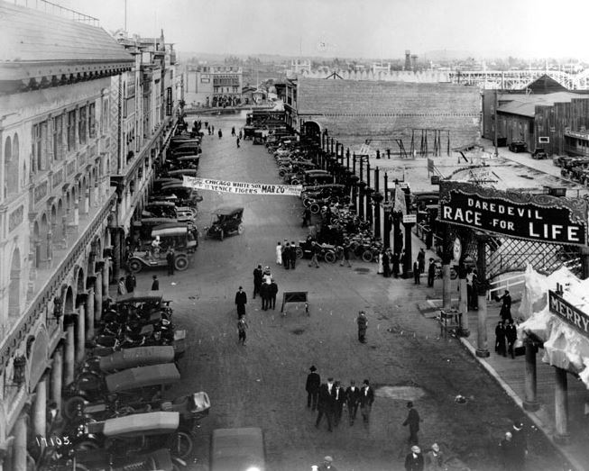  Looking inland from a roof at the end of Windward Avenue in Venice, one can see the street below and in the distance the lagoon and roller coaster. A banner across the street announces a baseball game between the Chicago White Sox and the Venice Tigers. On the right hand side of the street you can enter attractions such as the Daredevil Race for Life and the Merry Widow in 1913.