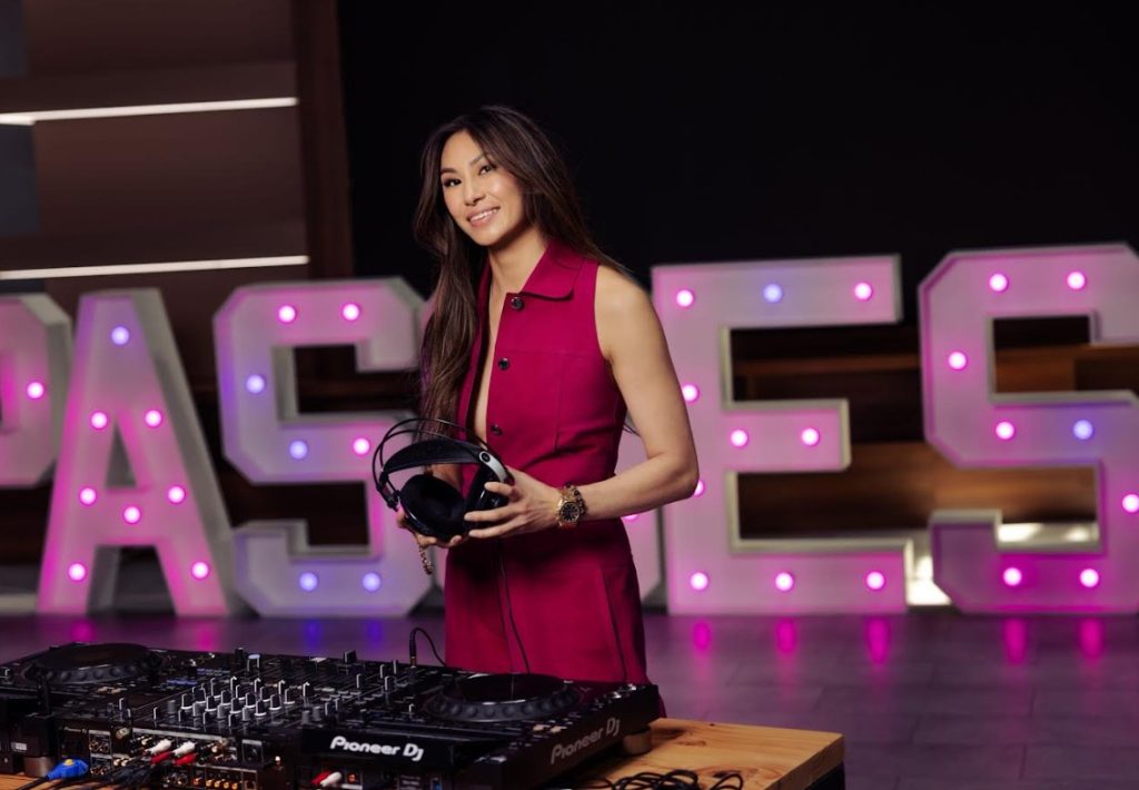 Lucy Guo holds a headset in front of a turntable in front of a neon sign that reads Passes.