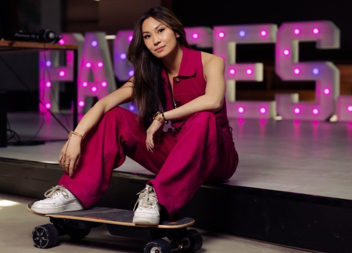Lucy Guo sits on a platform with her feet resting on a skateboard and a Passes neon sign in the background