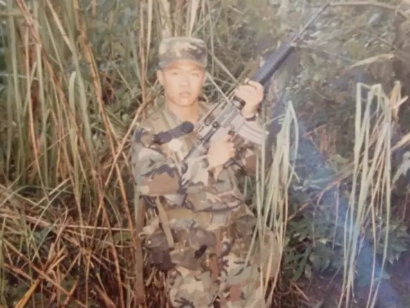 Sae Joon Park in military fatigues holding a rifle.